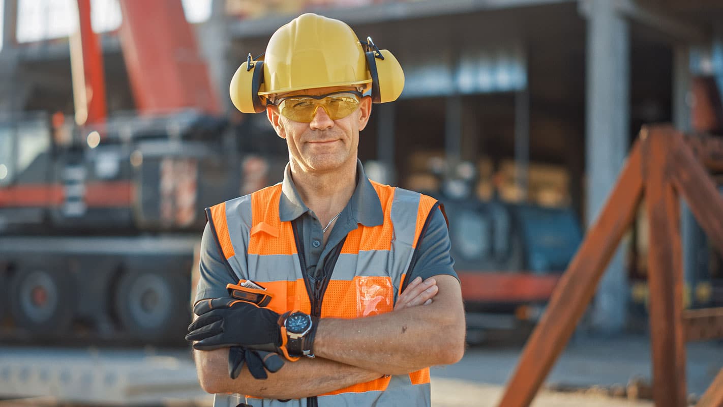 Portrait of Successful Builder / Worker / Contractor Wearing Hard Hat and Safety Vest Standing on a Commercial Building Construction Site, Crosses Arms Confidently. In the Background Crane Machinery.